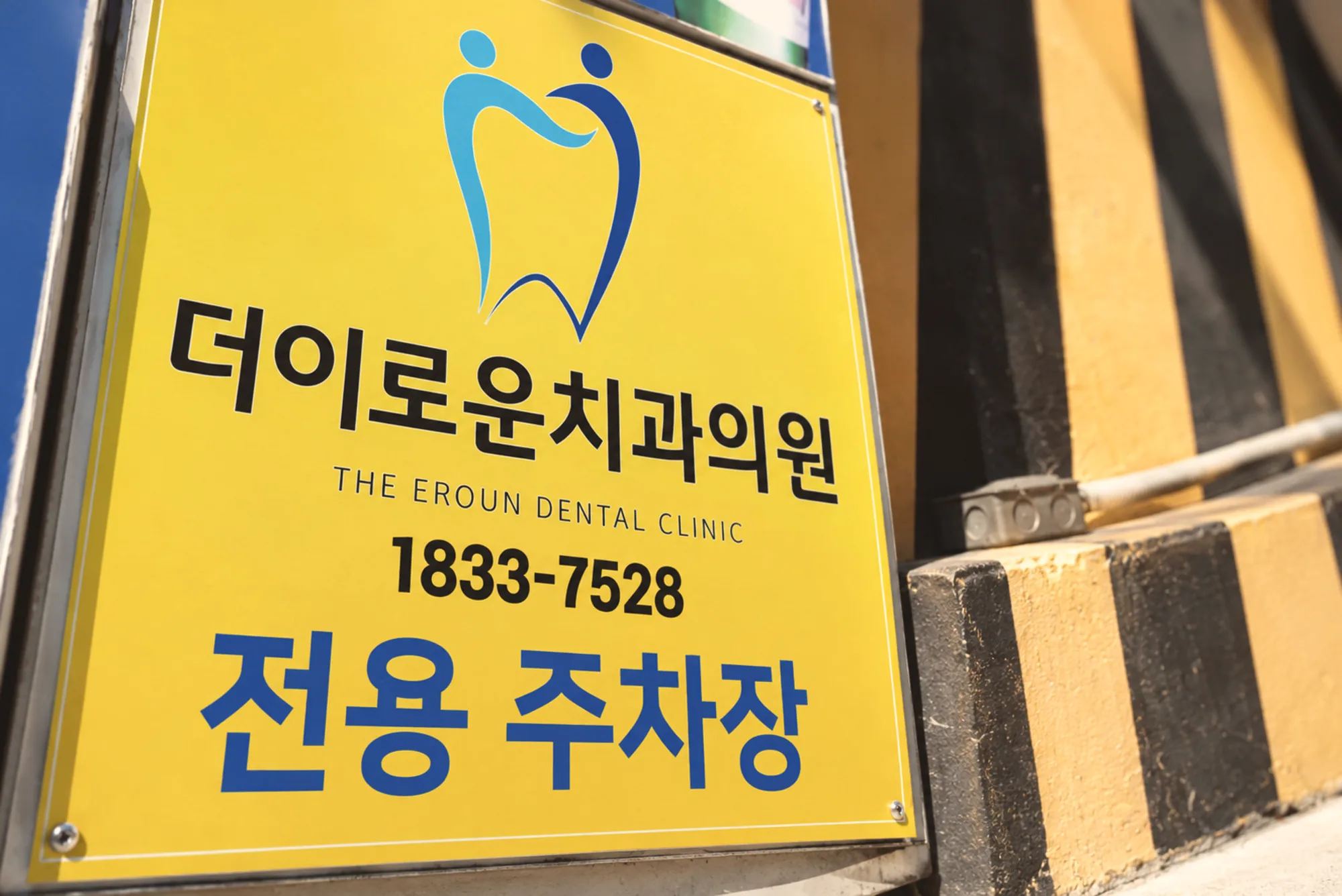 밝고 따뜻한 낮 자연광 아래 더이로운치과의원 외부 주차 안내 표지판과 검정·노랑 사선 벽을 담은 사진